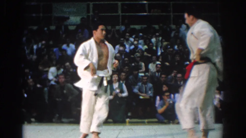 The competitors appear to reset during the 1966 West Covina judo match, one with an open gi jacket and another at frame right wearing a red belt.