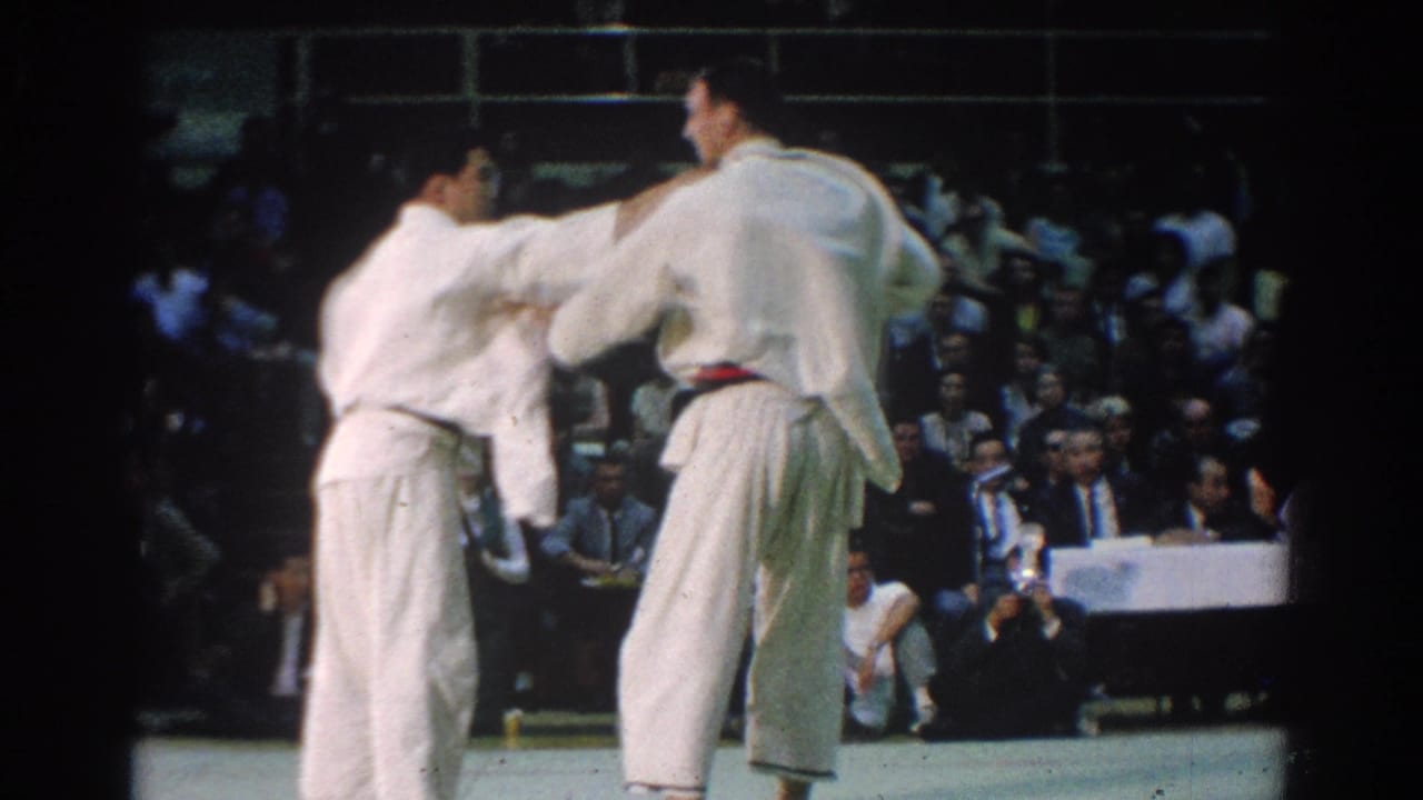 Two karate competitors in white gi grip each other on a mat before a seated audience.