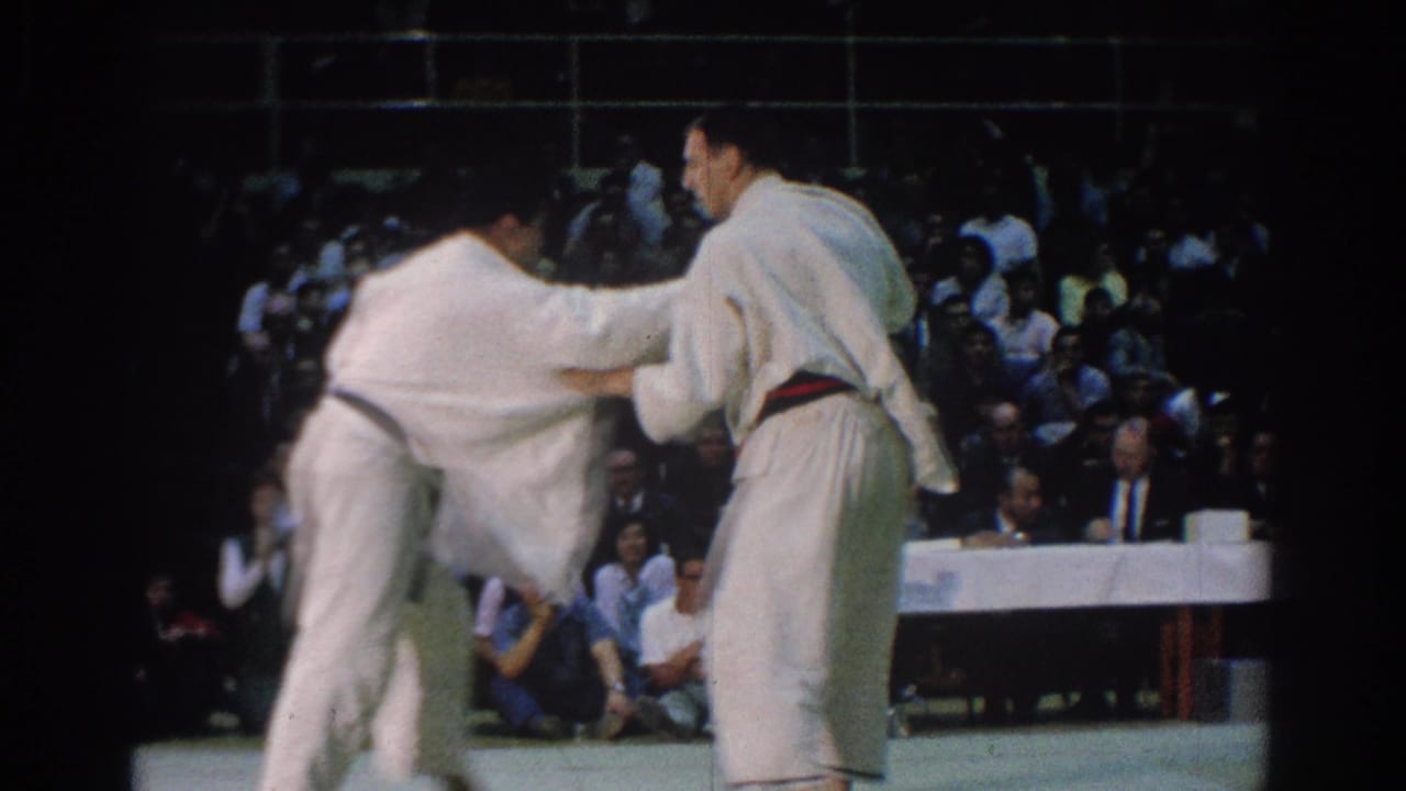 Two men in white gi grapple during the 1966 West Covina karate match, with a crowded spectator area behind them.