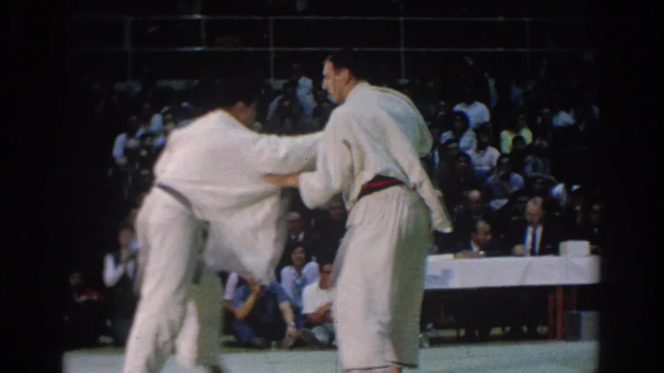 Two men in white gi grapple during the 1966 West Covina karate match, with a crowded spectator area behind them.