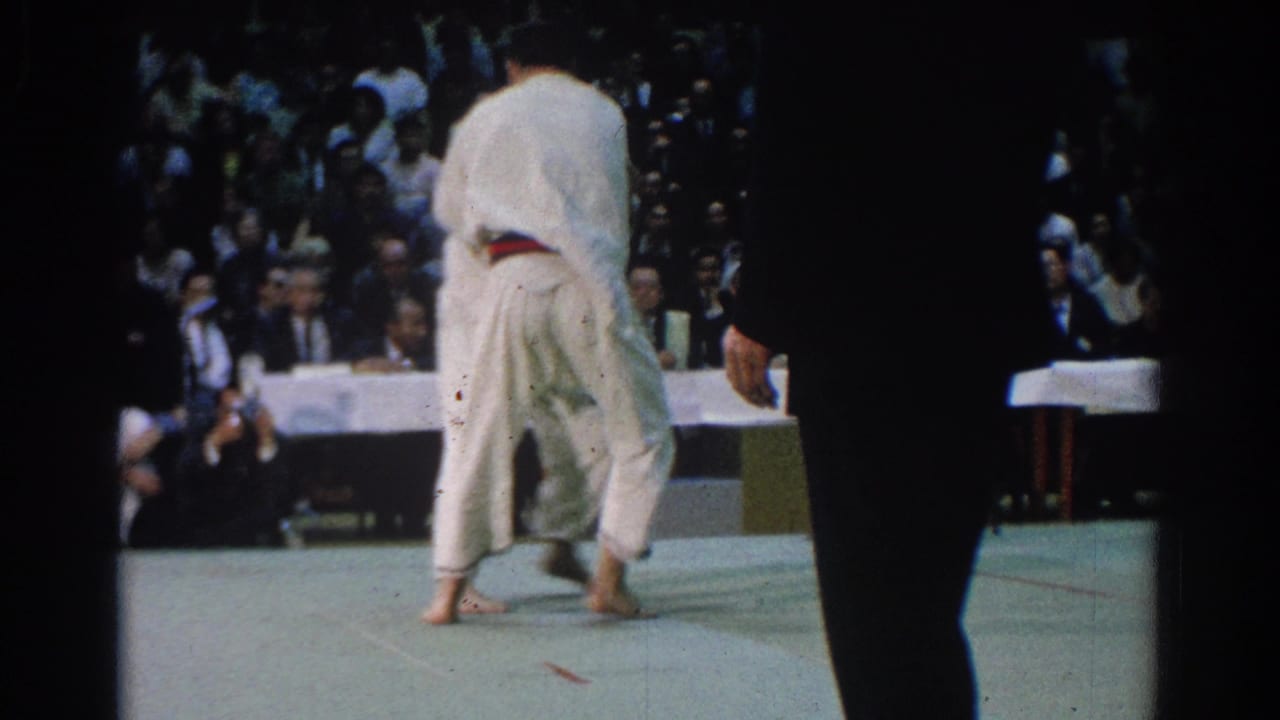 Two karate competitors in white gi grapple on a mat before seated spectators.