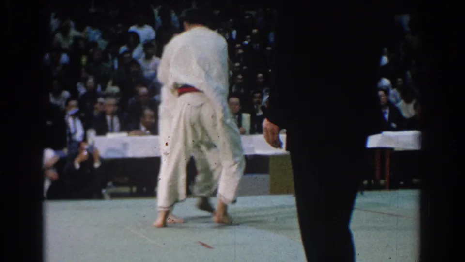 At a 1966 West Covina karate tournament, two men in white gi tussle on a light mat as judges and spectators watch.