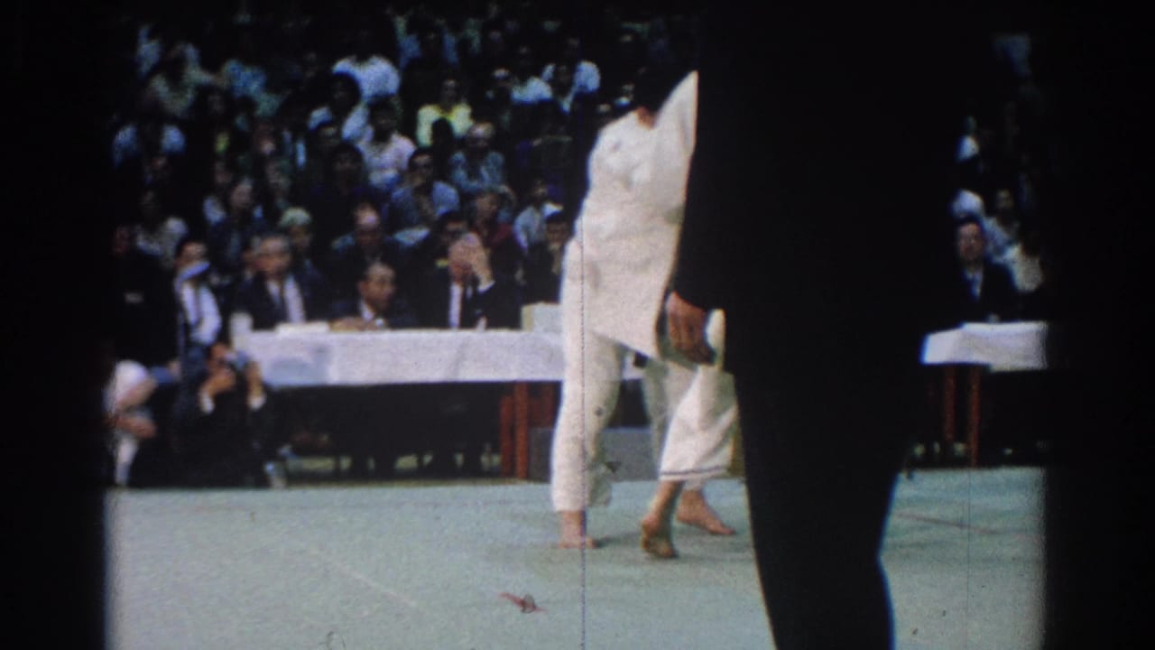 A karate competitor moves across the mat while a dark foreground figure partly blocks the view.