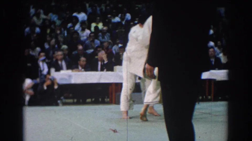 During the West Covina match, one white-clad competitor steps across the mat while the audience and judges remain visible behind him.