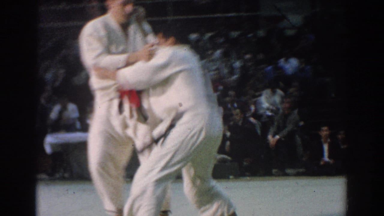 A white-clad competitor bends low while holding his opponent during the West Covina karate match, with spectators blurred in the background.