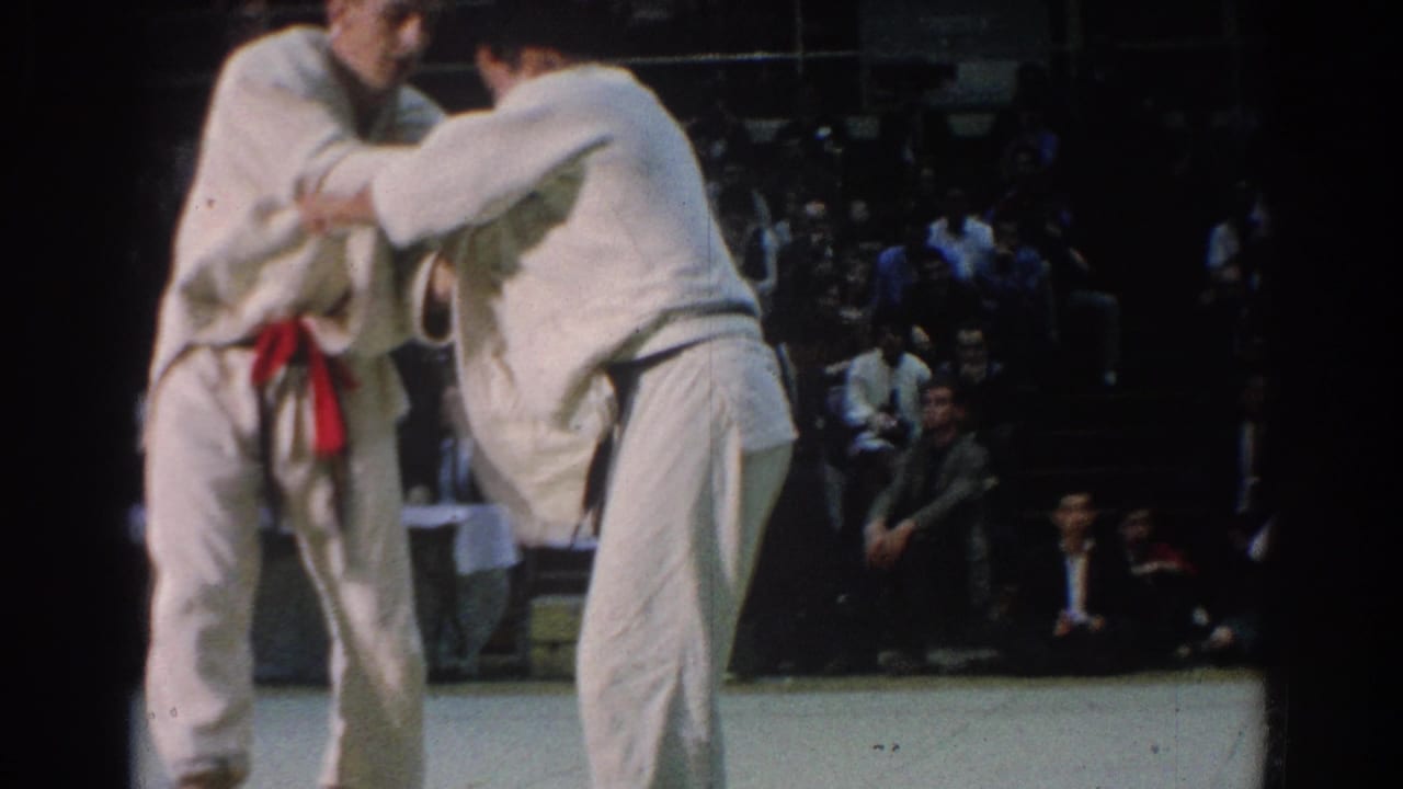 Two judokas in white gi grapple on an indoor mat with spectators in dark bleachers behind them.