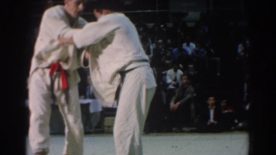 In 1966 West Covina, two adult male judokas in white gi grip each other during an indoor match as spectators watch from bleachers.