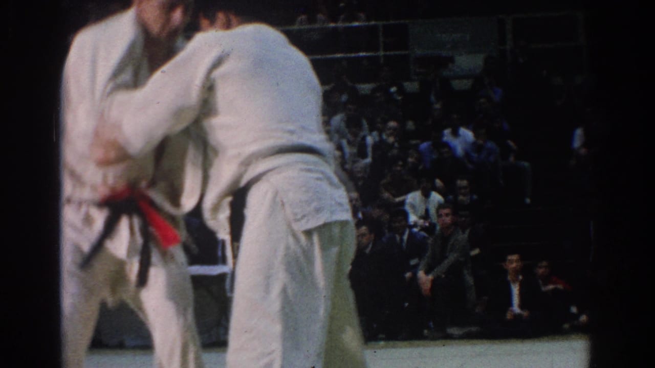 Two white-uniformed judokas continue grappling near the mat edge as seated spectators watch.