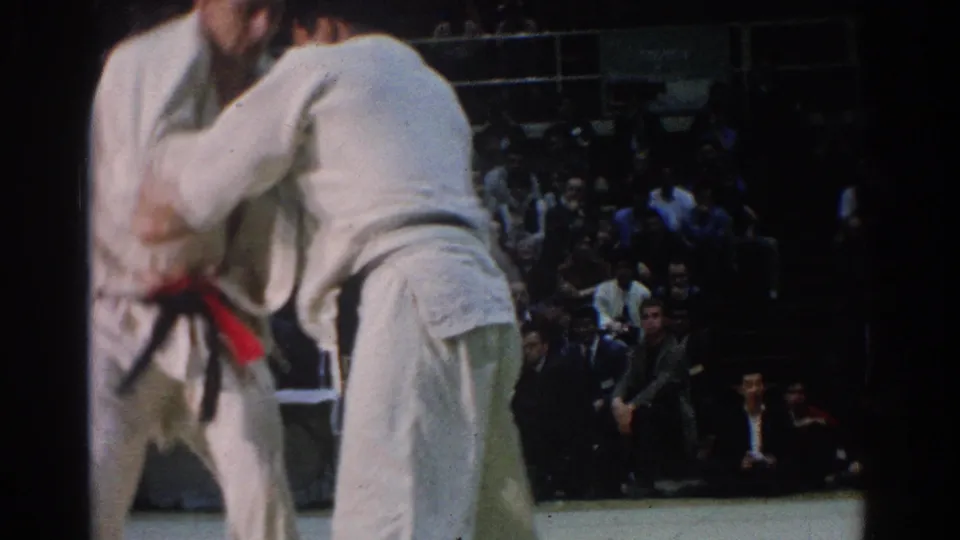 The 1966 West Covina judo match continues with the competitors locked in close grips beneath bright indoor arena lighting.