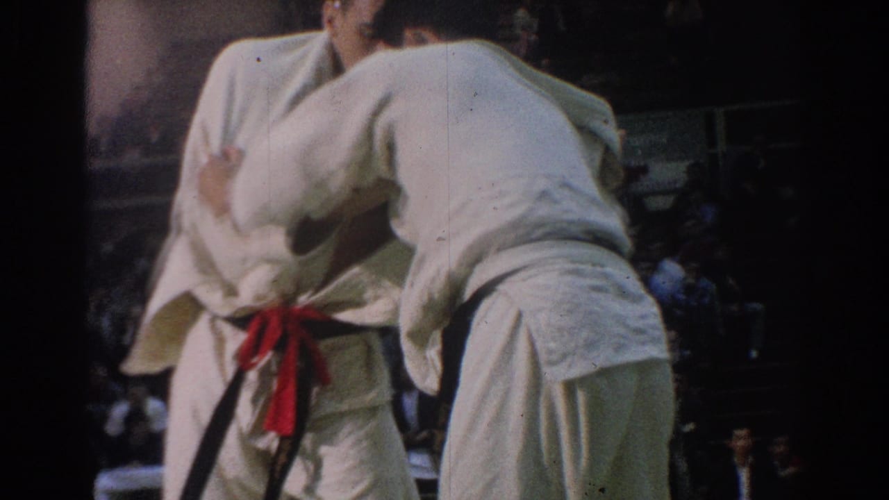 Two men in white martial arts uniforms grapple in front of seated spectators.