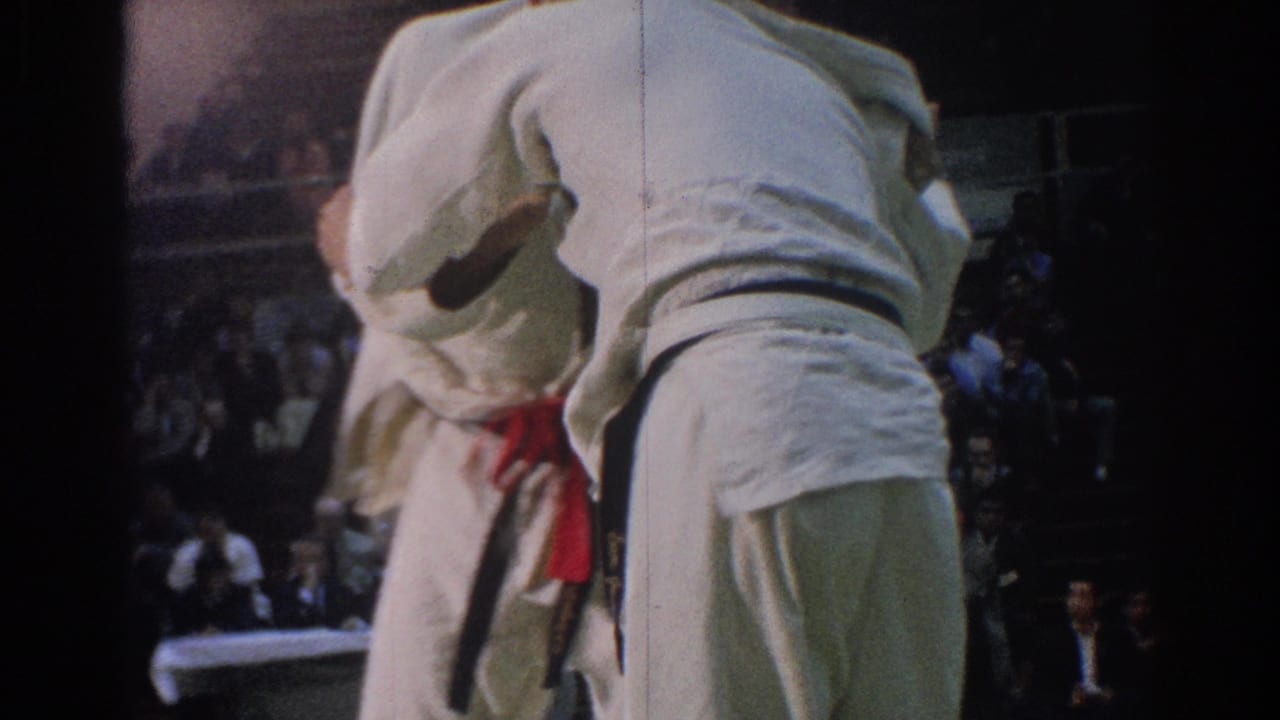 Two male competitors in white gi lean into each other during a West Covina martial arts match, with a dark spectator area behind them.
