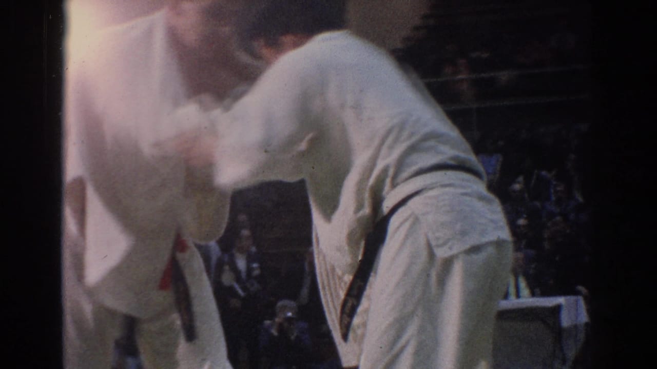 Two adult karate competitors lean into a close hold with spectators behind them.