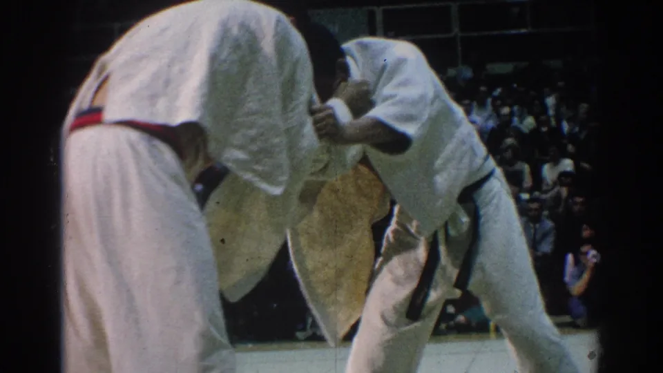 A 1966 West Covina karate match frame shows one adult competitor bent forward while another maintains a close grip near the crowd.