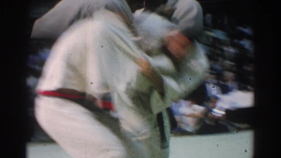 1966 West Covina 8mm frame showing two judo athletes in white gis grappling at close range, with motion blur and spectators behind them.