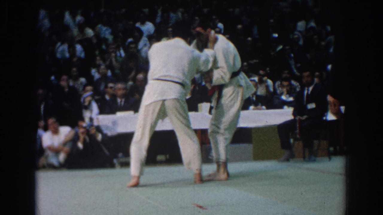 1966 West Covina frame with two athletes in white judo uniforms squared up on the mat while spectators and officials watch from behind.