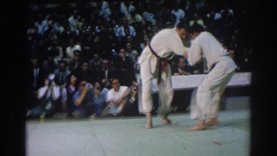 1966 West Covina 8mm frame showing two judo athletes gripping sleeves and lapels, with photographers seated near the mat and audience behind.
