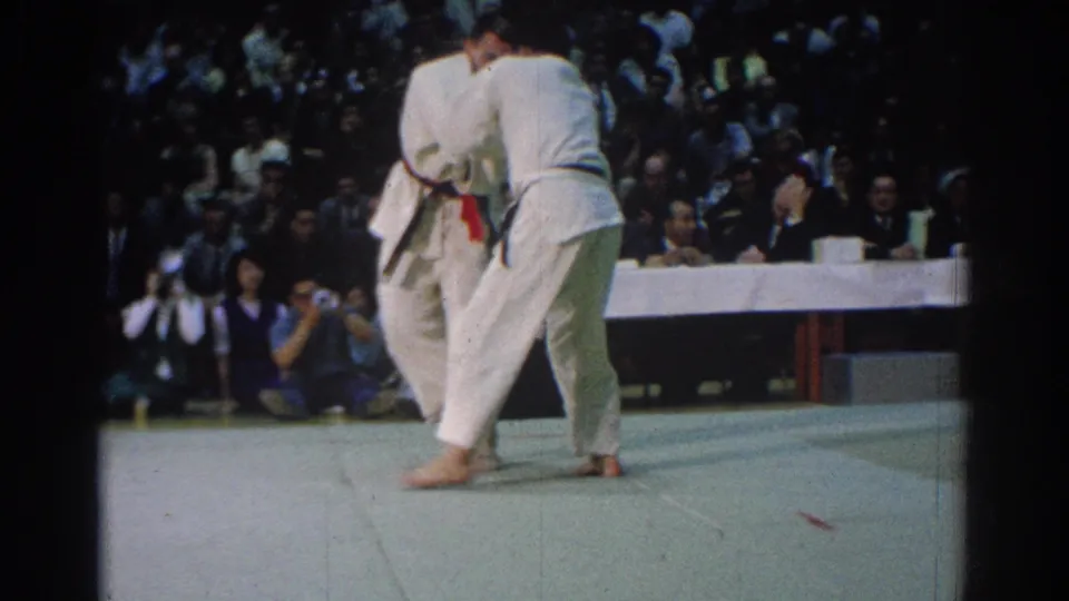 1966 West Covina frame of two judo competitors in white gis locked in a standing grapple, with spectators and photographers visible behind them.