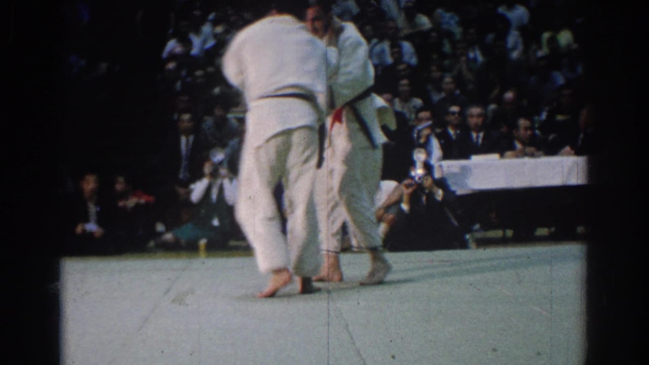 Two judokas in white uniforms grapple on a mat before a seated crowd.