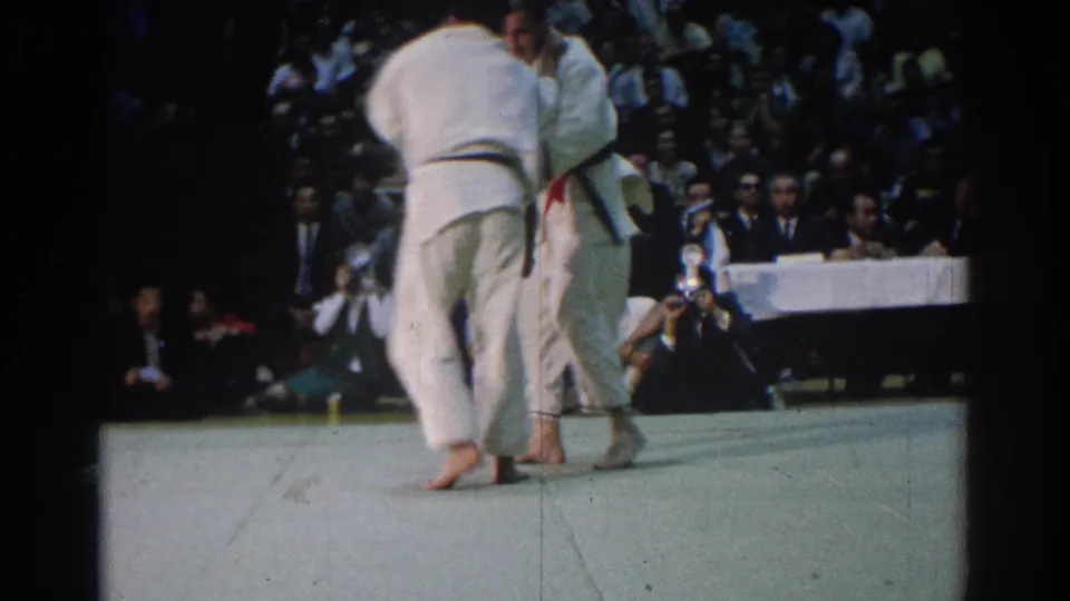 At a 1966 West Covina judo competition, two black-belt judokas grip each other near the edge of a light mat as spectators watch.