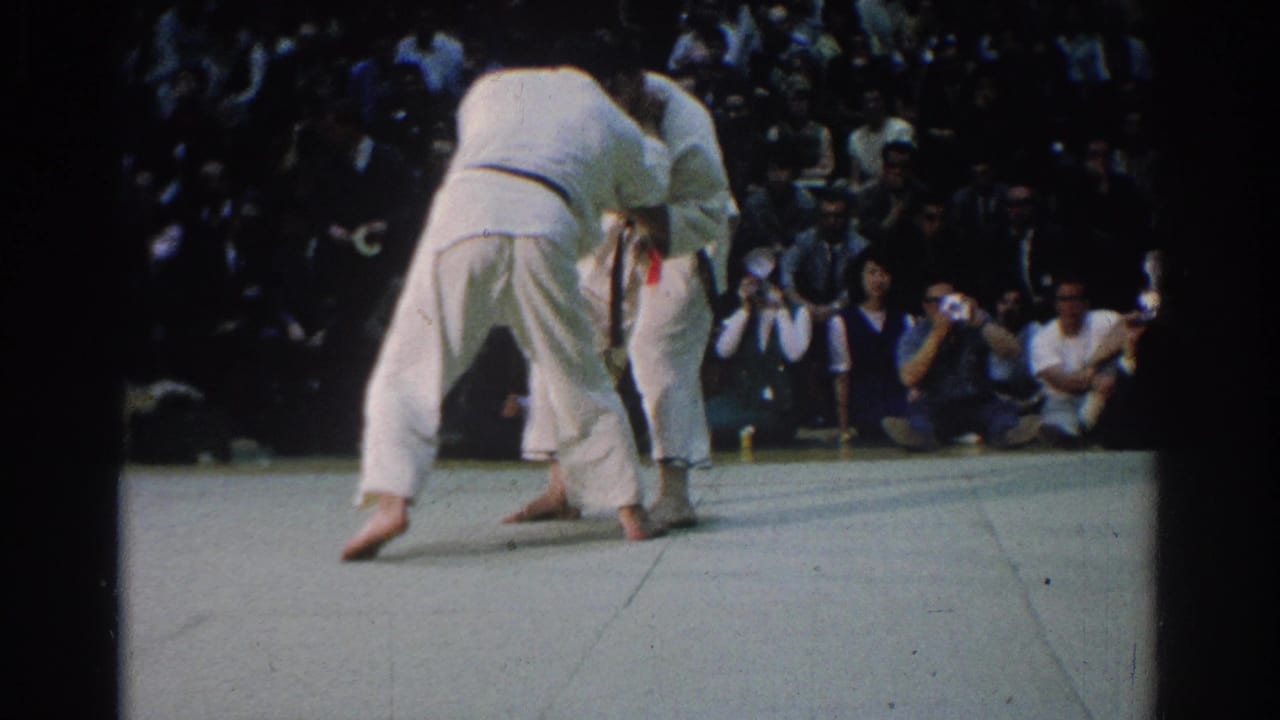 The West Covina match continues as the judokas bend forward in close grips, with photographers and audience members visible beyond the mat.
