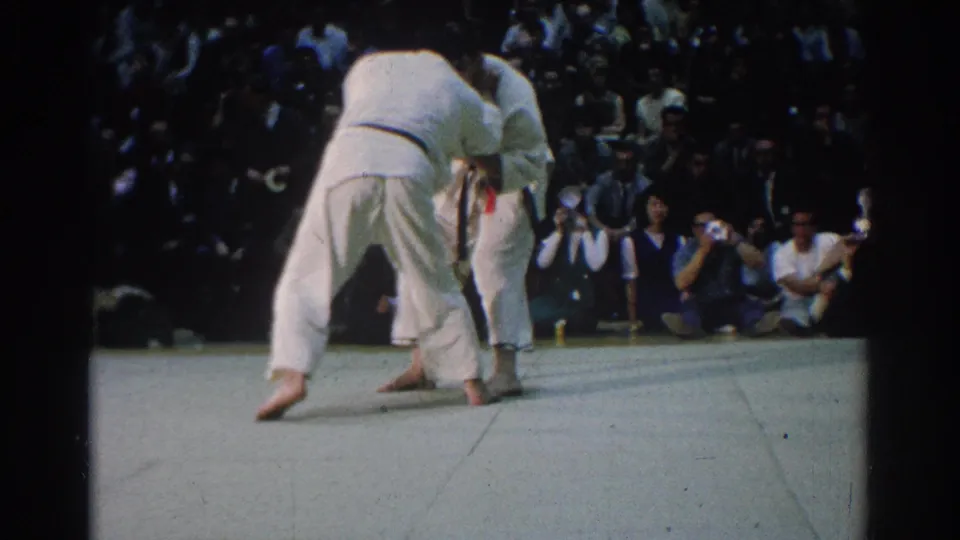 The West Covina match continues as the judokas bend forward in close grips, with photographers and audience members visible beyond the mat.