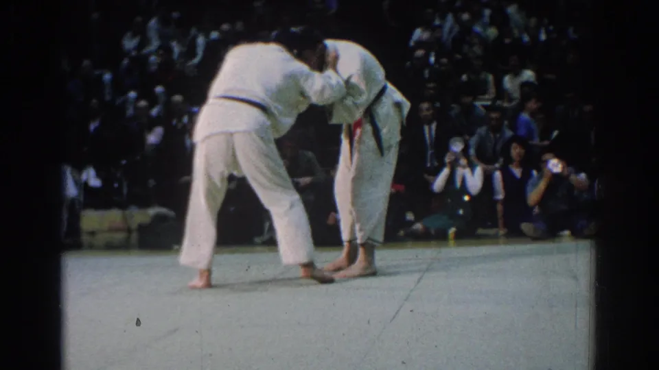 One judoka bends low while holding his opponent's sleeve or lapel during the 1966 judo match, with spectators and photographers behind the mat.