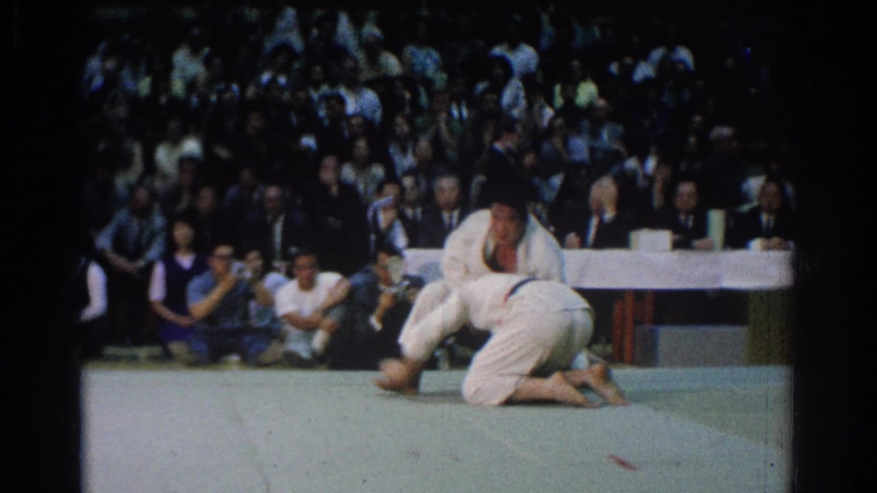 Two judokas grapple low on a mat as spectators watch from behind a table.