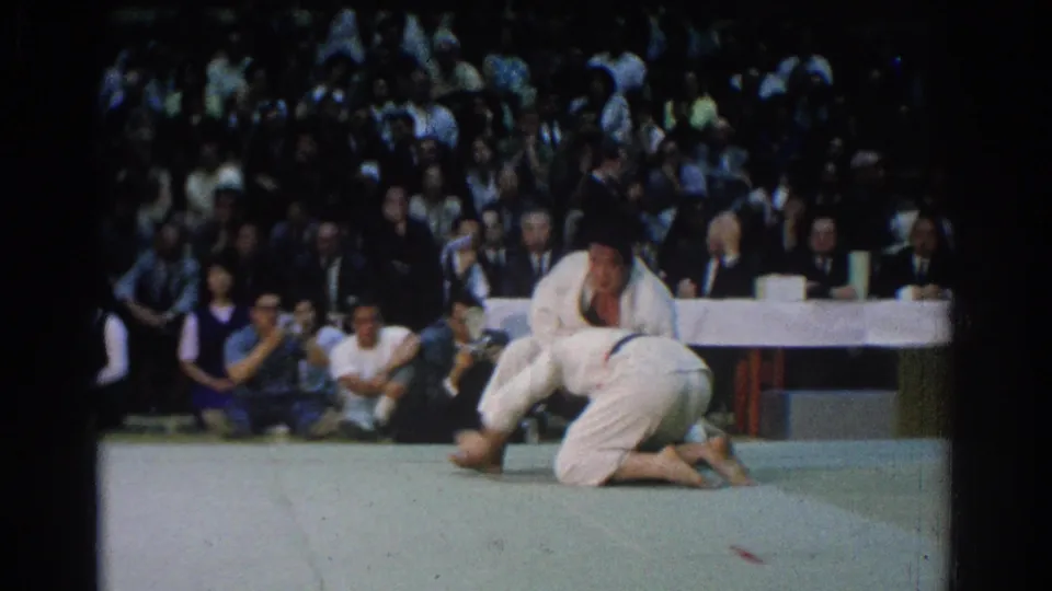 In 1966 West Covina footage, two judokas in white gi grapple near the mat while a seated audience and officials watch behind them.