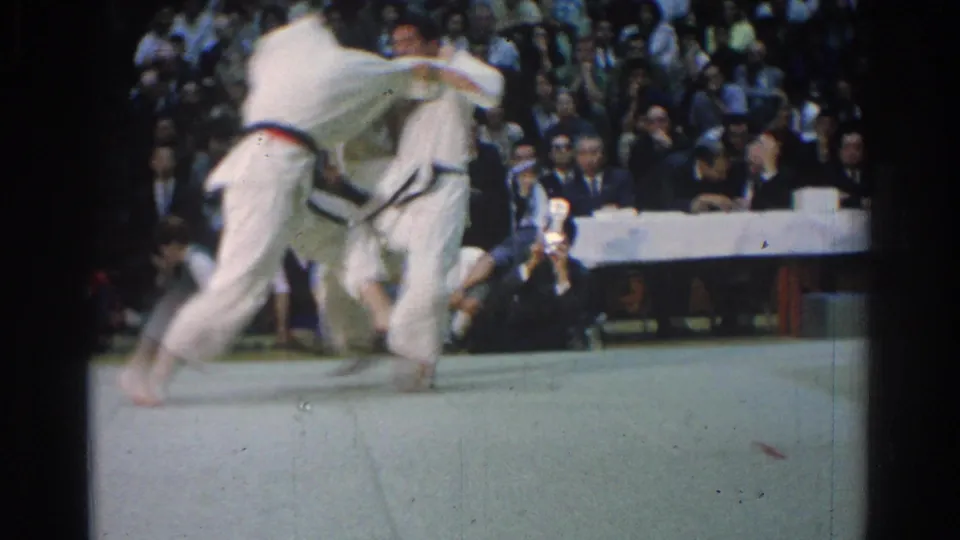 Two white-gi competitors blur through a standing judo throw on the mat, with spectators and officials visible in the indoor arena.