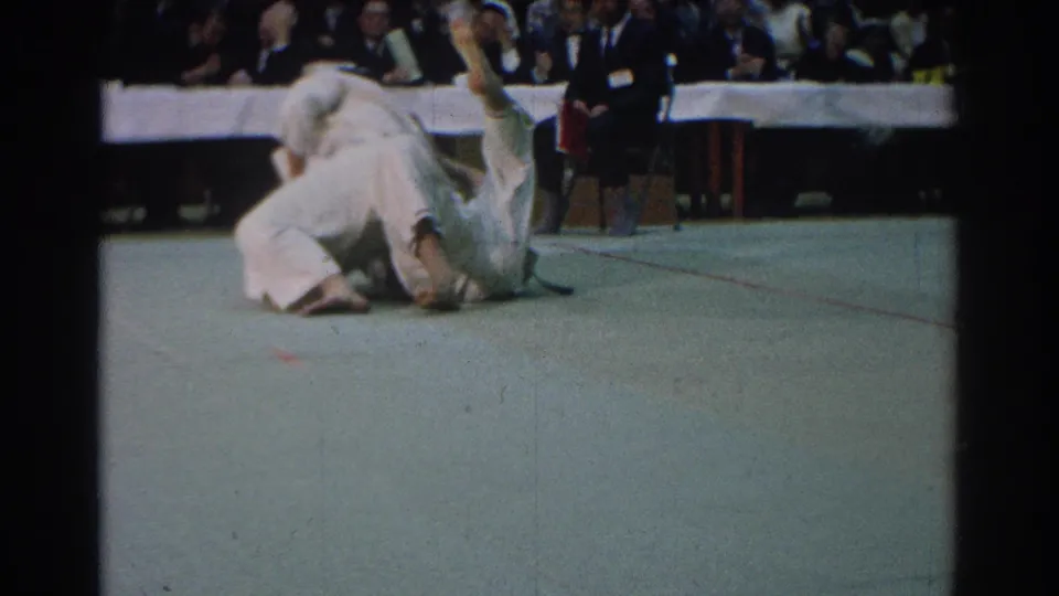 Two competitors continue ground action on the judo mat while a referee stands close by and the indoor audience looks on.