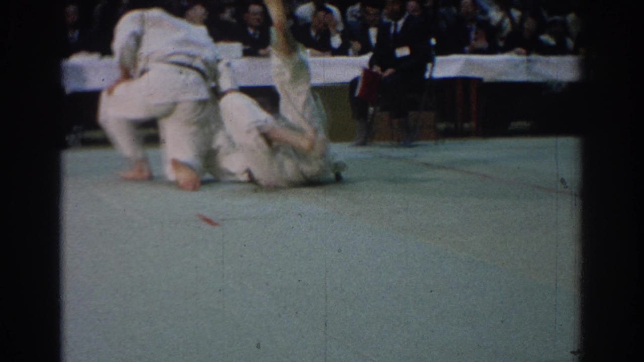 In 1966 West Covina footage, two judo competitors in white gi are on a green mat as spectators watch from behind tables.