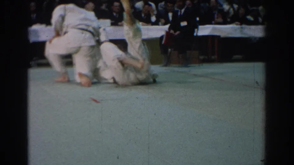 In 1966 West Covina footage, two judo competitors in white gi are on a green mat as spectators watch from behind tables.