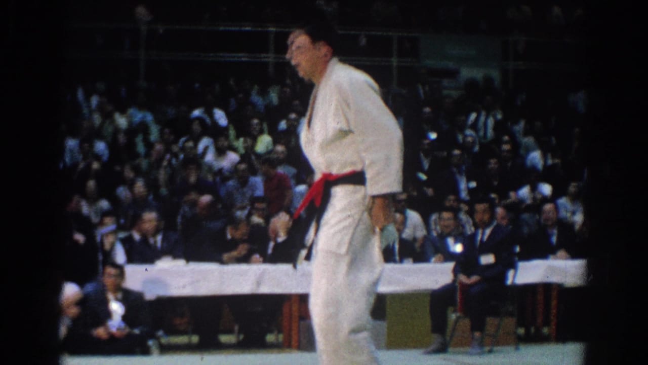A red-belt judo competitor in a white gi crosses the mat during the 1966 West Covina tournament, with spectators in the background.