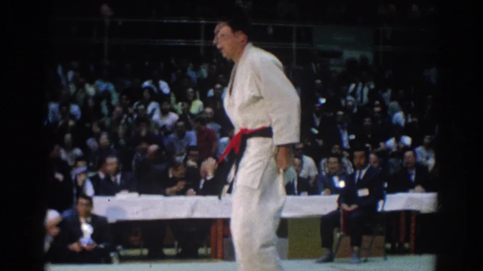 A red-belt judo competitor in a white gi crosses the mat during the 1966 West Covina tournament, with spectators in the background.