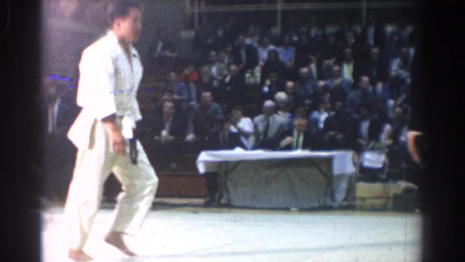 A judo competitor in a white gi moves across the mat area in West Covina, with a seated audience and officials table behind him.