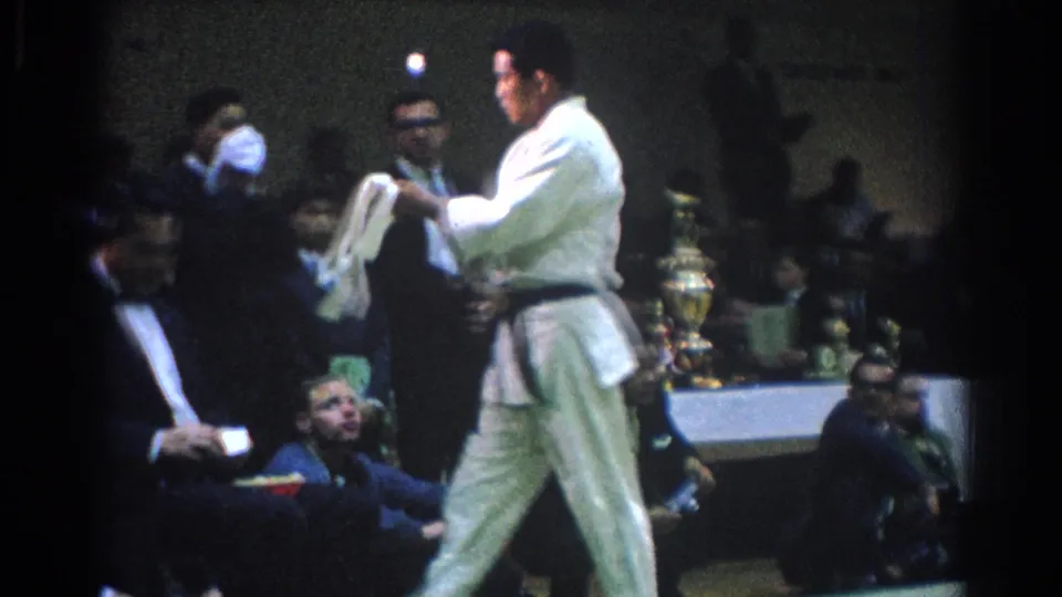 A judo competitor stands near officials and trophies at the 1966 West Covina event, with audience members visible around the mat.