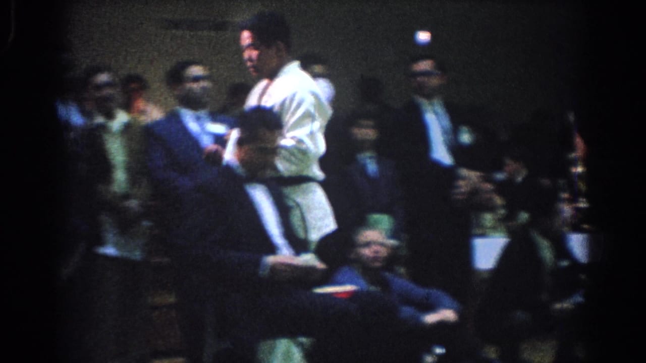1966 West Covina footage shows a martial artist in a white gi standing among seated officials and spectators in a crowded indoor arena.
