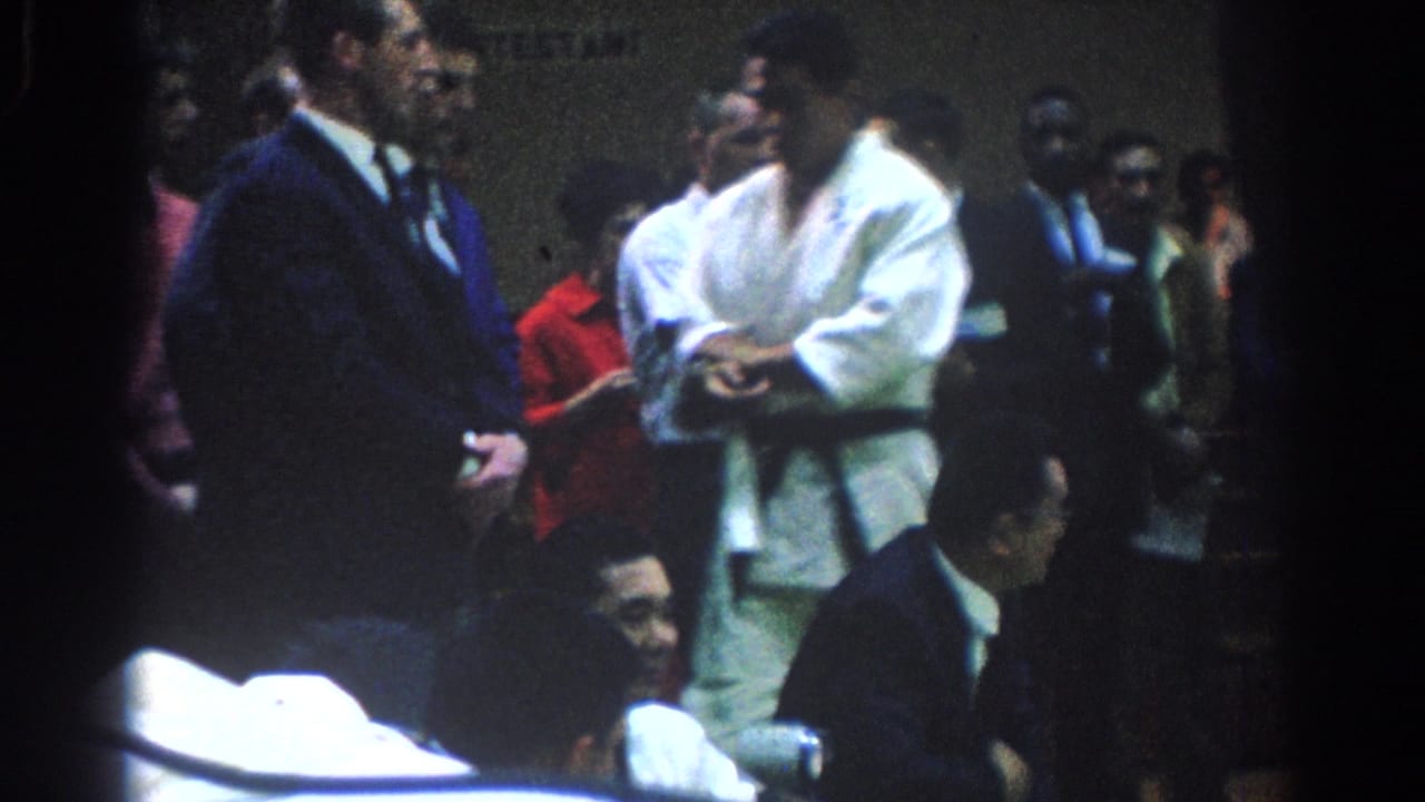 Man in white gi stands beside a suited man while spectators sit and watch behind them.