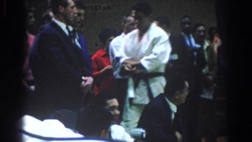 In the West Covina martial arts setting, a competitor in a white gi stands near a suited official with spectators gathered around.