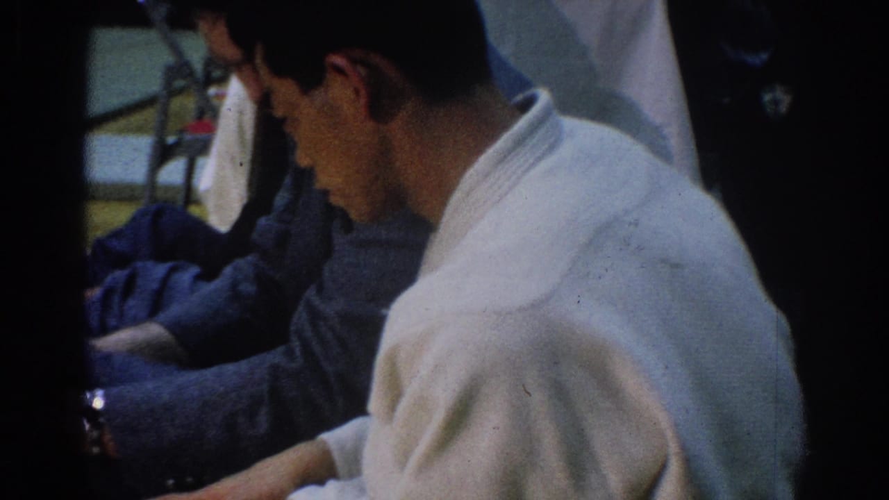 Man in a white robe sits beside suited adults in a dim interior.