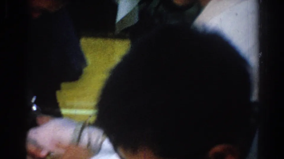 1966 West Covina home-movie frame with blurred foreground heads and a bare foot being handled in a dim room.