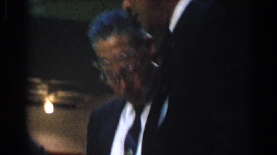 1966 West Covina frame showing an older man with glasses in a dark suit, partly blurred in low interior light.