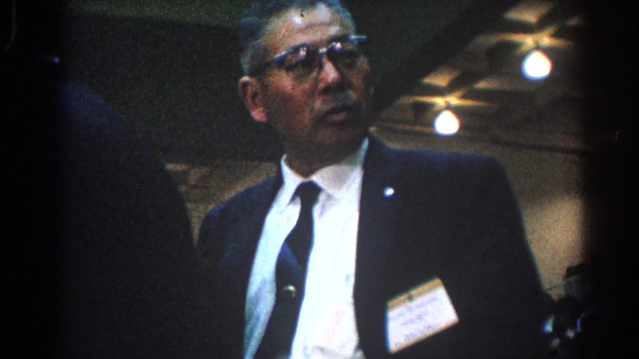 Suited official with glasses and a name badge seen indoors near event seating.