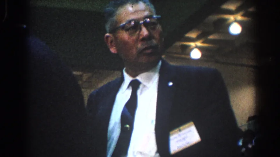 In West Covina, California, 1966, a suited official with glasses and a name badge stands or moves into view inside an indoor event space.