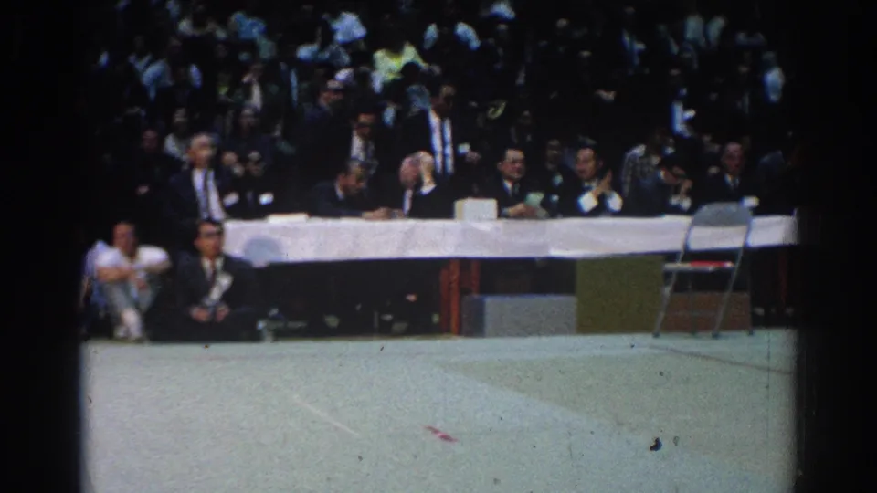 The camera frames the judges table, seated spectators, and open mat area during the West Covina sporting event.