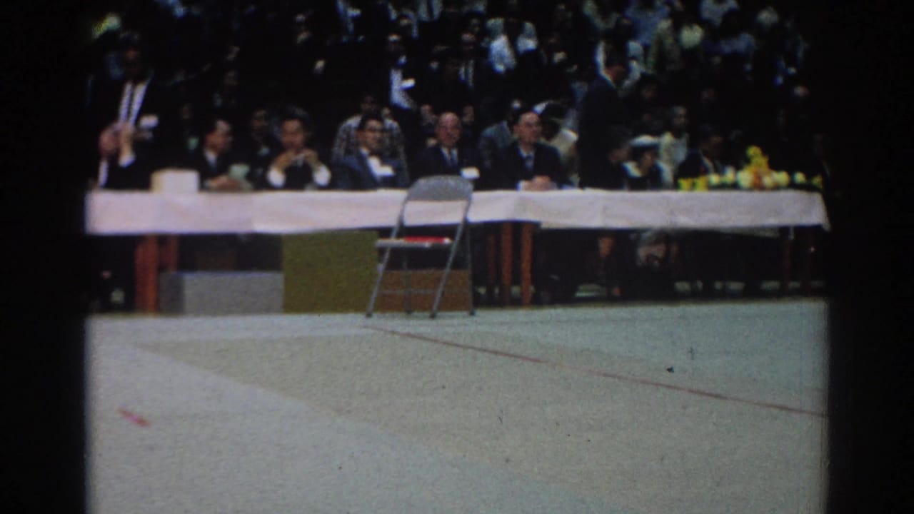 A 1966 West Covina indoor event view shows suited men at a long table, a crowd behind them, and an empty chair on the floor.