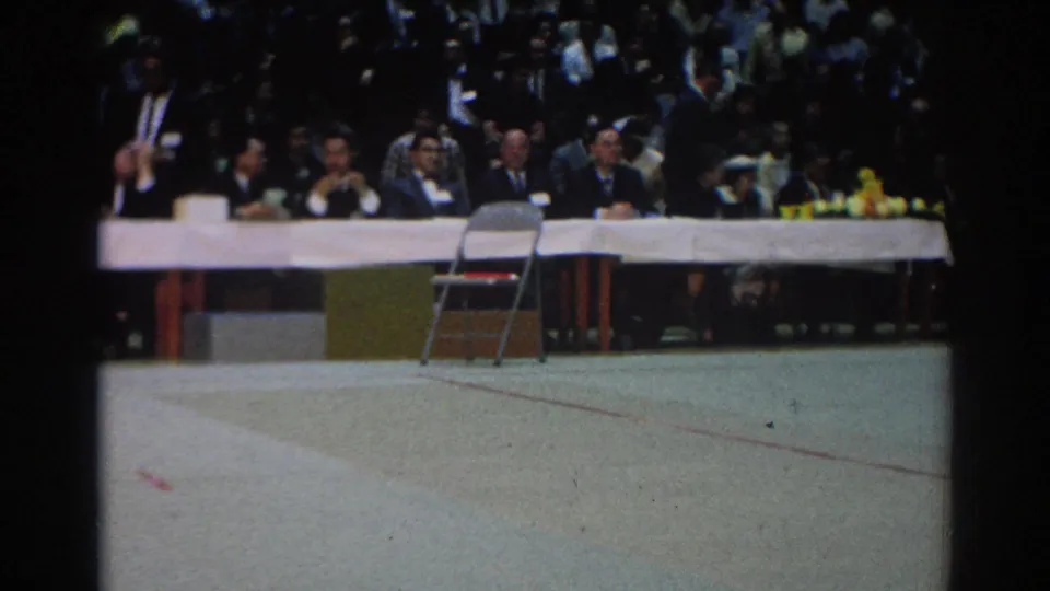 A 1966 West Covina indoor event view shows suited men at a long table, a crowd behind them, and an empty chair on the floor.