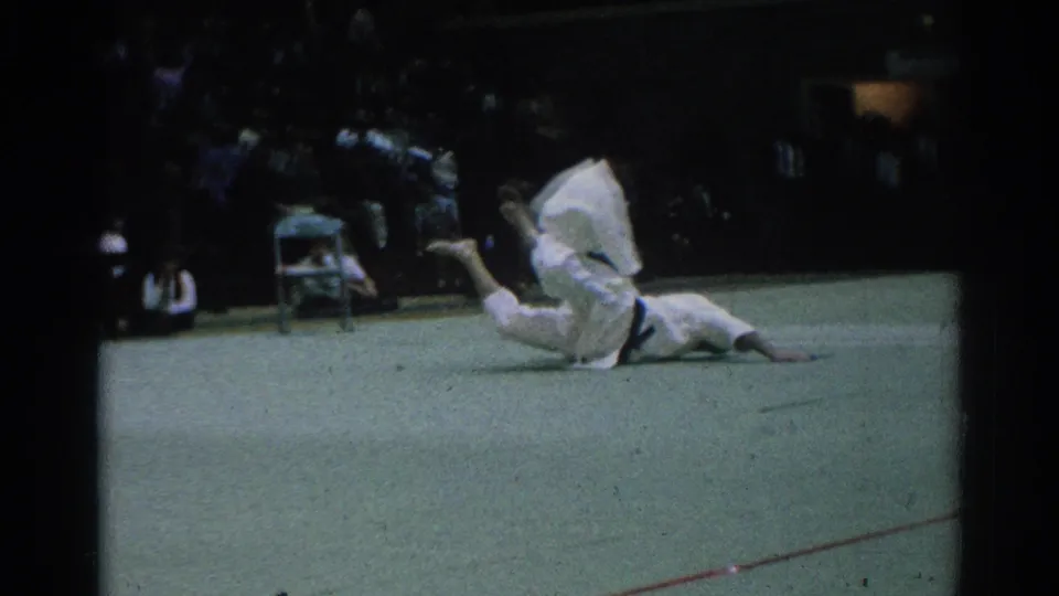 A karate participant in a white uniform appears on the floor during the 1966 West Covina indoor event, with spectators behind.