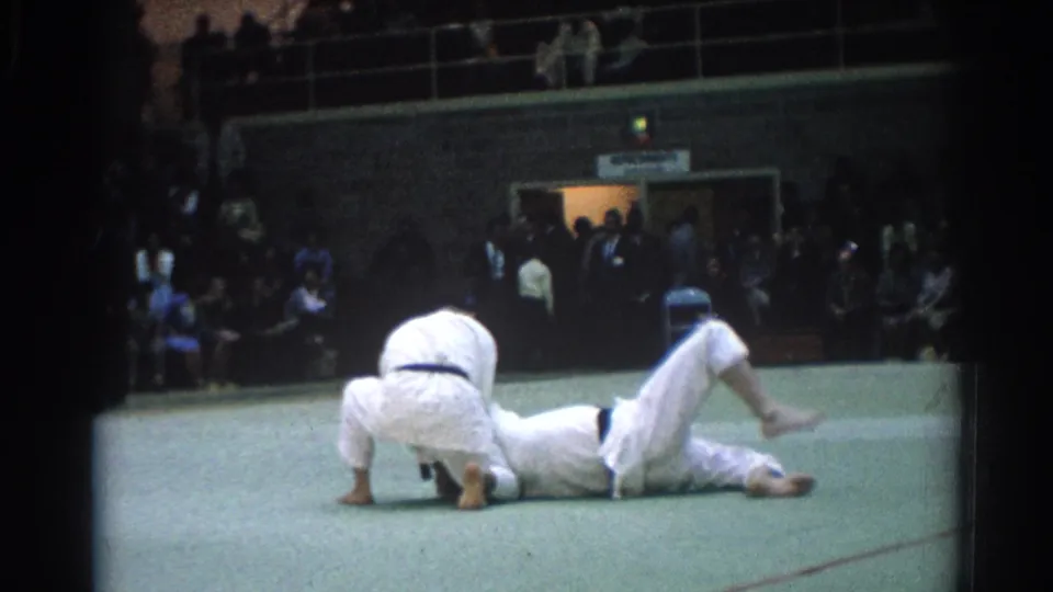 Two competitors continue grappling on the green mat after a takedown during a public martial arts match.