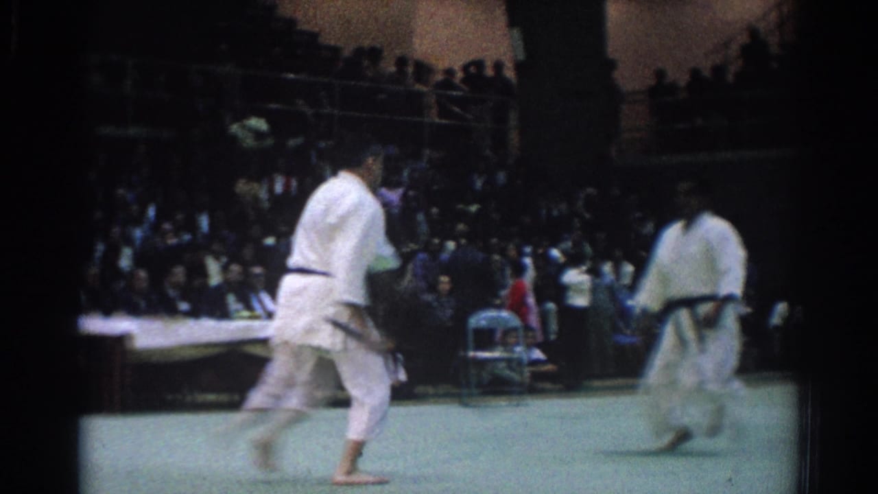 Two judo fighters in white gi face each other on a green mat before a seated crowd.